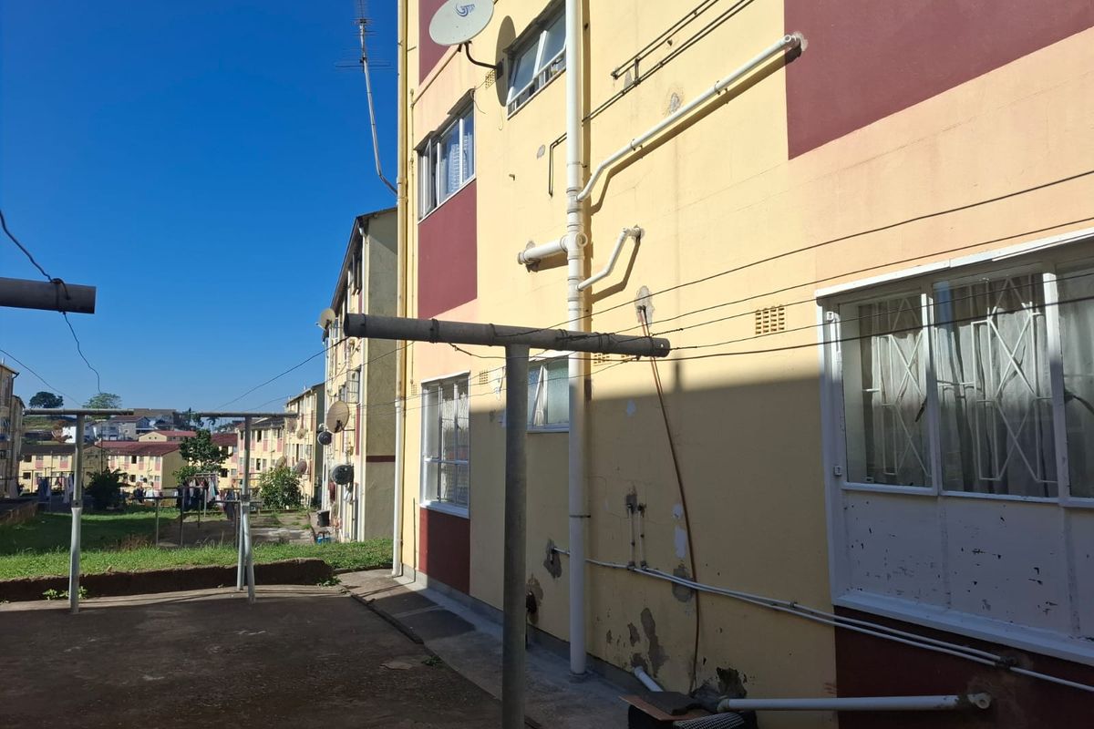 Rear exterior view of an apartment building showing concrete paving, external plumbing, and weathered wall finishes with security bars on windows.