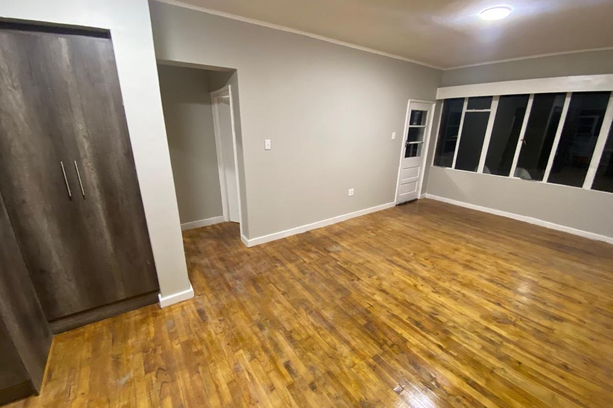 An empty living room featuring original wooden parquet flooring, a built-in cupboard, and large windows providing natural light.