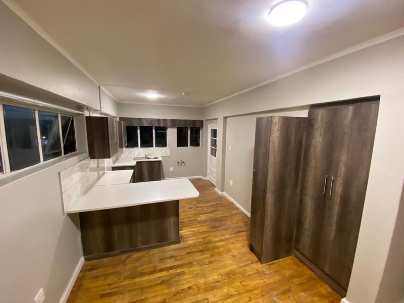 A clean, modern kitchen featuring dark wood-look cabinetry, white countertops, tiled backsplash, and warm laminate flooring in an open-plan layout.