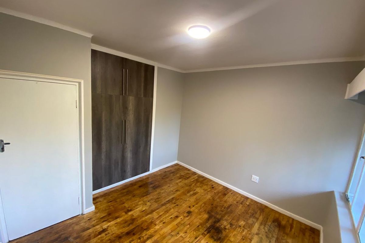 A bright, empty bedroom featuring polished wooden flooring, neutral grey walls, and modern built-in cupboards with dark wood-grain finish.