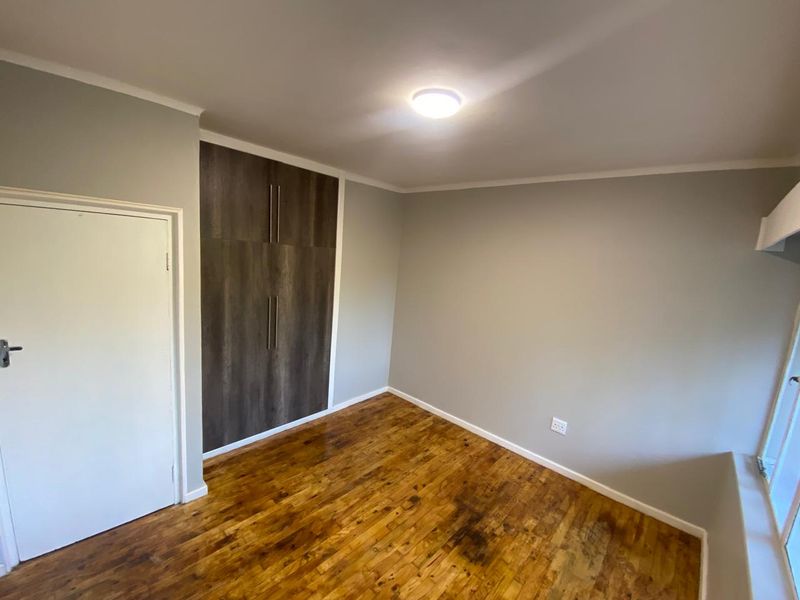 A bright, empty bedroom featuring polished wooden flooring, neutral grey walls, and modern built-in cupboards with dark wood-grain finish.
