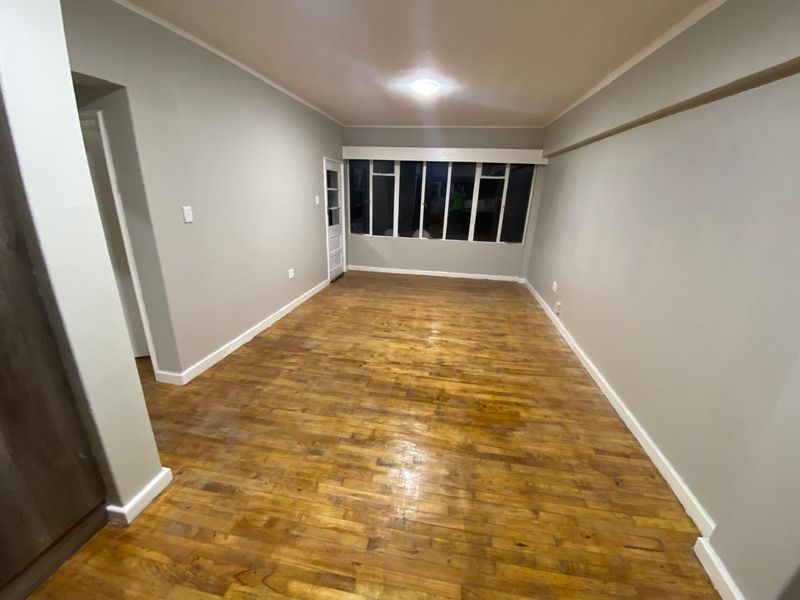 A spacious, empty living area featuring polished parquet flooring, neutral wall tones, and large windows providing natural light.