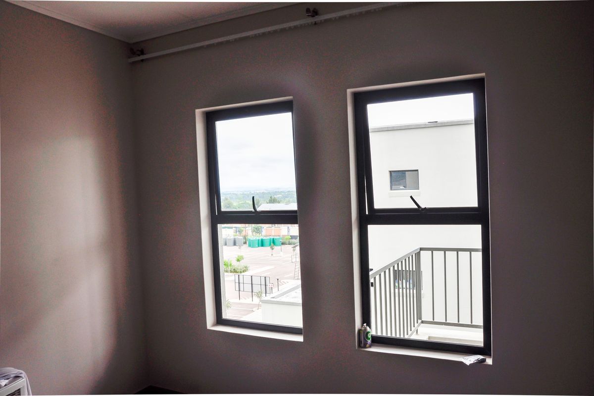 A bright bedroom featuring two modern dark-framed windows with views of the surrounding complex and exterior balcony.