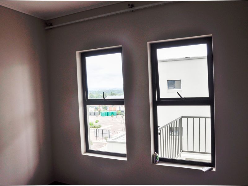 A bright bedroom featuring two modern dark-framed windows with views of the surrounding complex and exterior balcony.