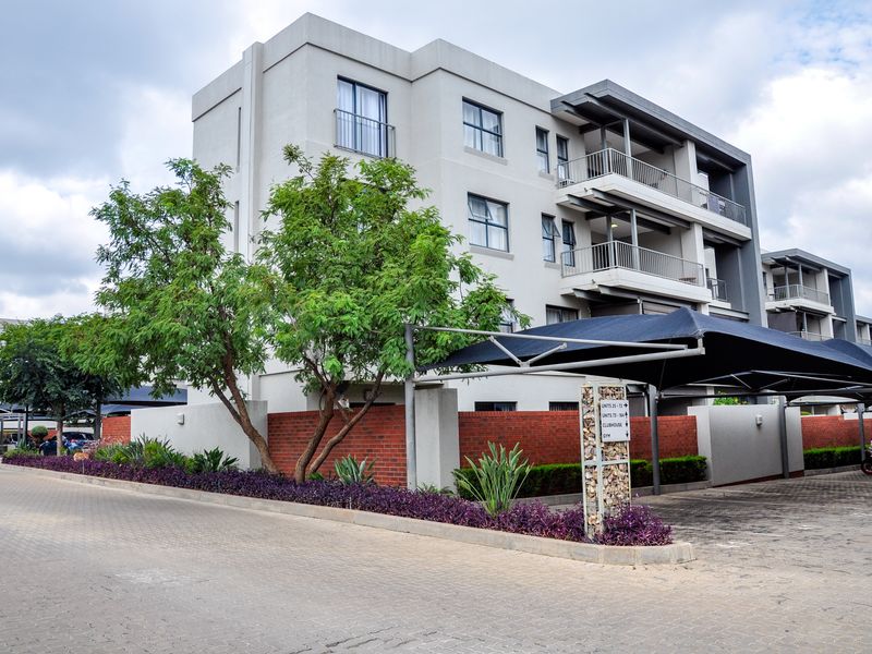 Exterior view of a modern apartment complex featuring multi-level balconies, paved parking areas with shade sails, and landscaped garden beds.