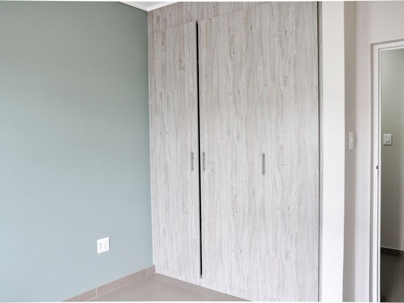 A bedroom featuring built-in wood-look cabinetry and a neutral accent wall, with tiled flooring and a doorway leading to an adjacent room.