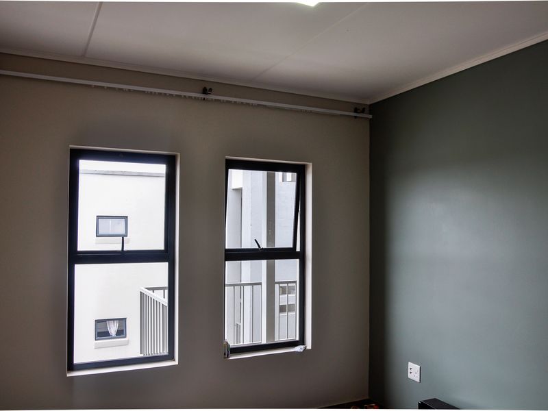 A bright bedroom featuring dual aluminium-framed windows, a neutral color palette with a dark accent wall, and integrated electrical outlets.