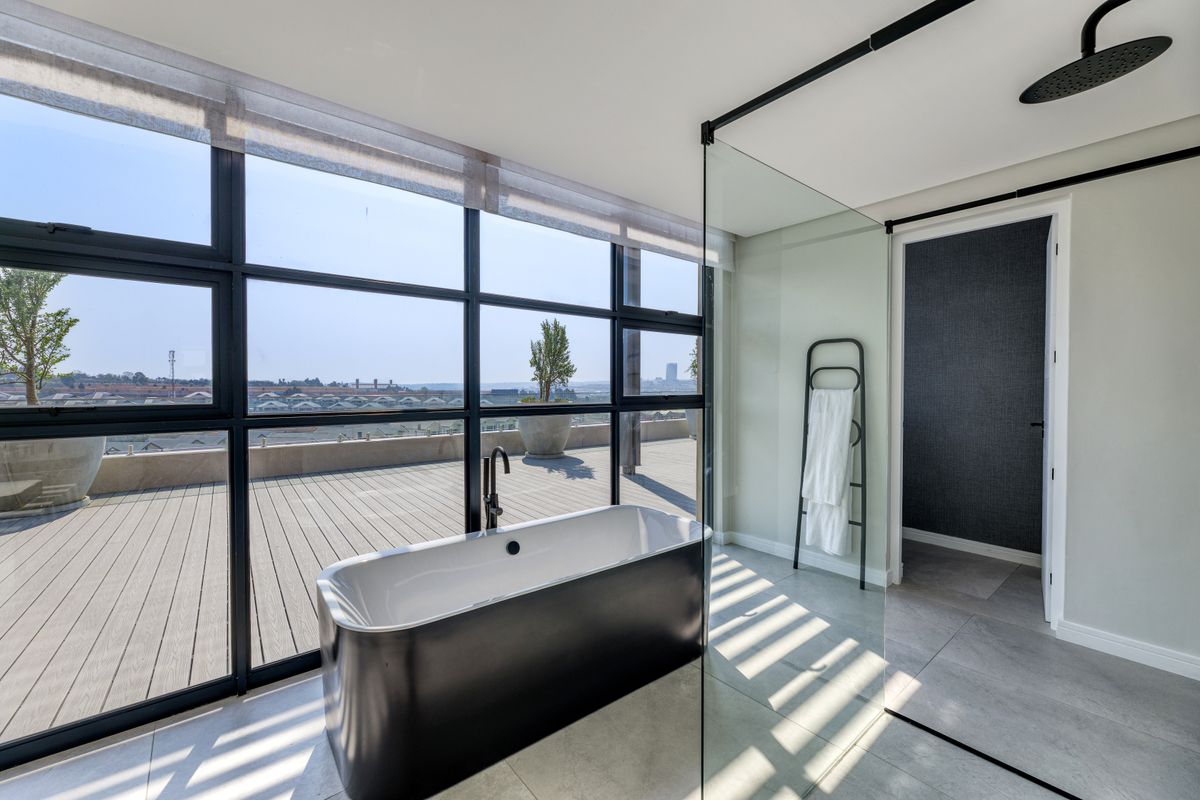 Modern penthouse bathroom featuring a freestanding bathtub, glass-enclosed rainfall shower, and floor-to-ceiling windows opening onto a spacious wooden deck with panoramic views.