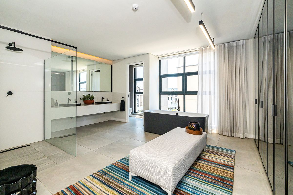 Modern open-plan bathroom featuring a glass-enclosed walk-in shower, dual vanity, and a freestanding bathtub with large windows and floor-to-ceiling wardrobe storage.