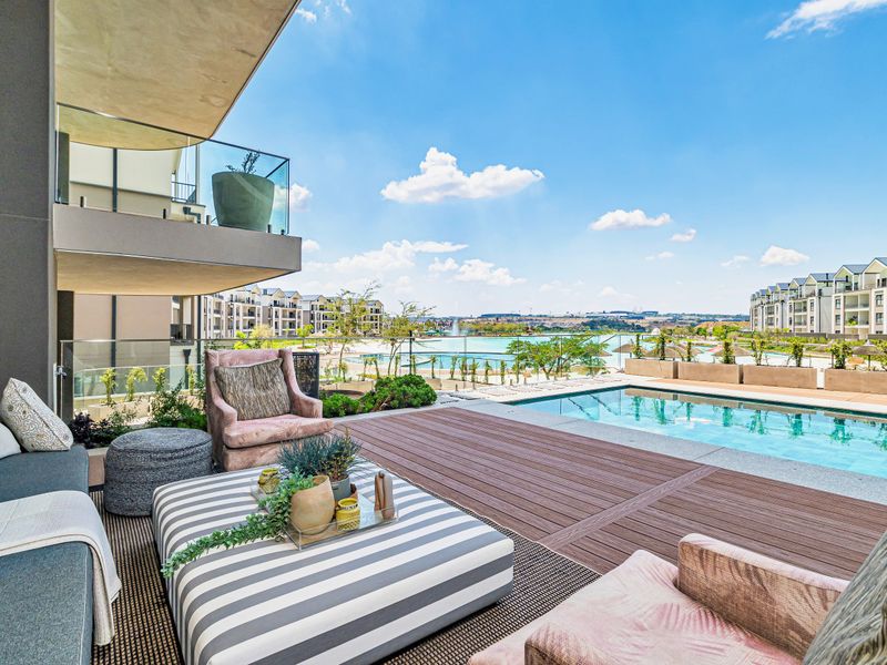 A spacious covered patio featuring composite decking, overlooking a private swimming pool and a scenic lagoon view within a modern apartment complex.