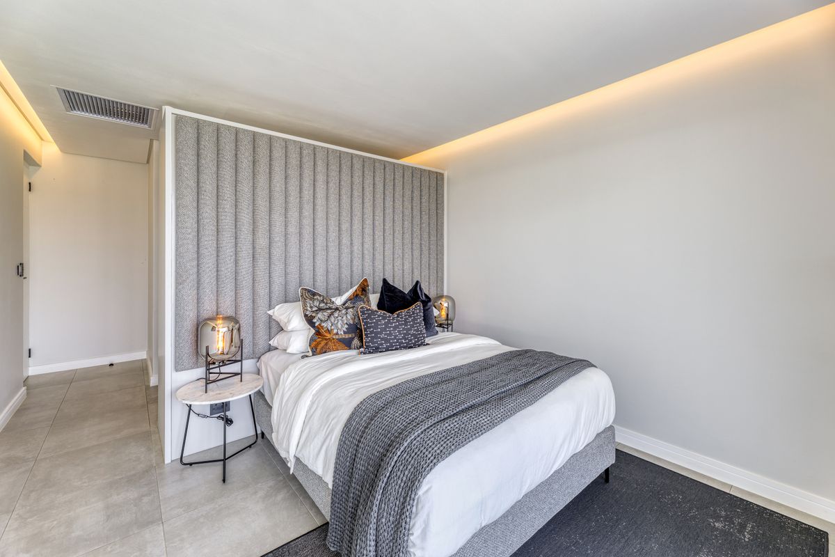 A modern bedroom featuring a custom upholstered headboard wall, recessed ceiling lighting, and neutral tiled flooring in a contemporary apartment setting.