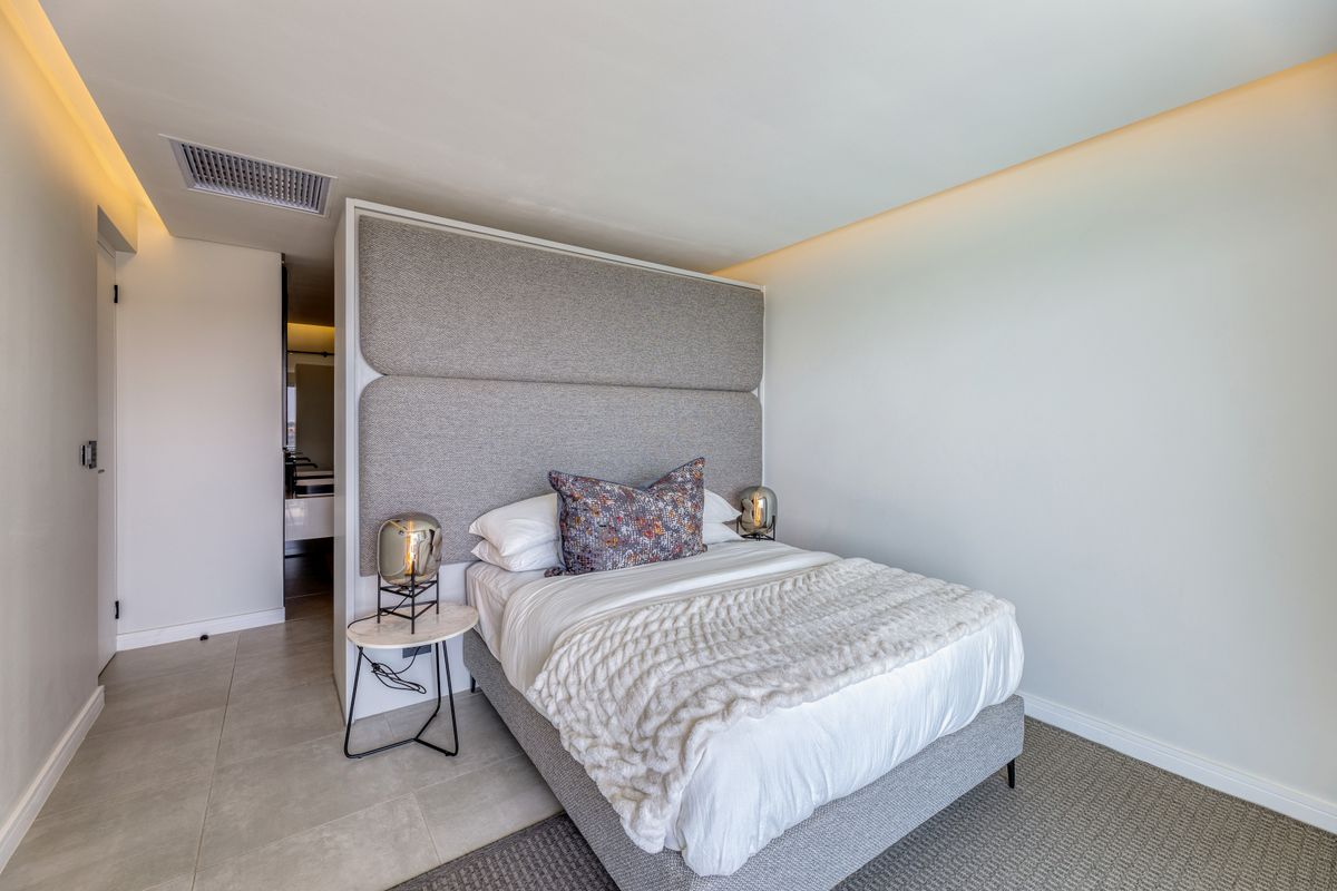 A modern bedroom featuring a built-in upholstered headboard, recessed ceiling lighting, and integrated air conditioning vent, with neutral finishes and clean lines.