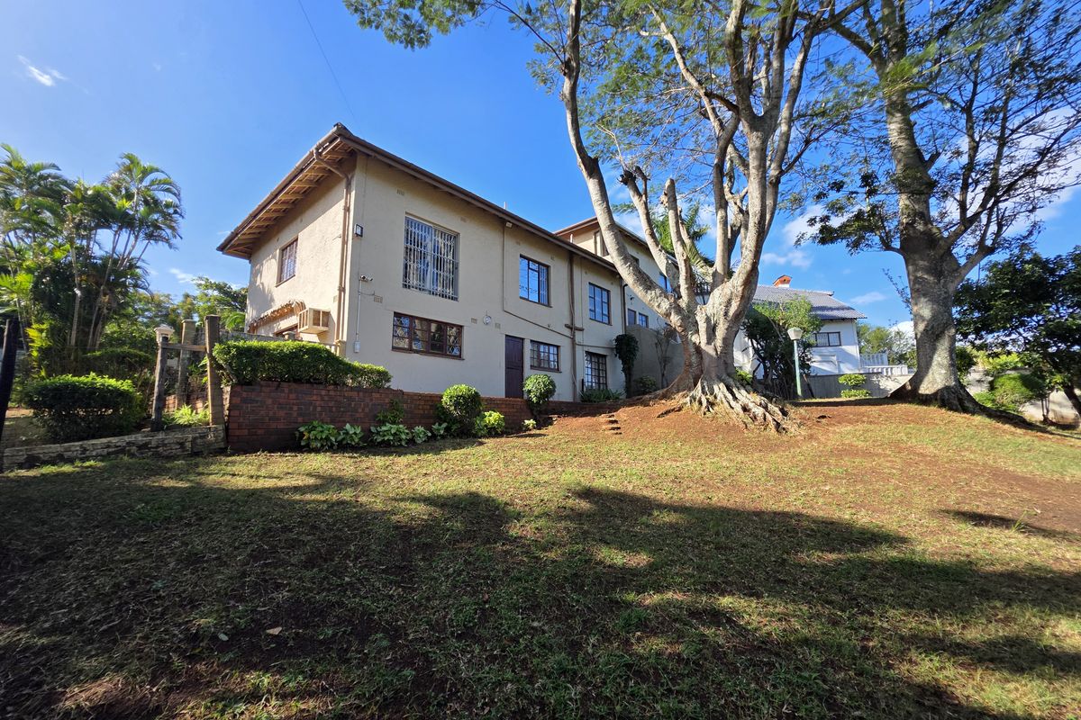 Rear view of the property featuring a multi-story building facade, brick retaining wall, and established garden with mature trees.