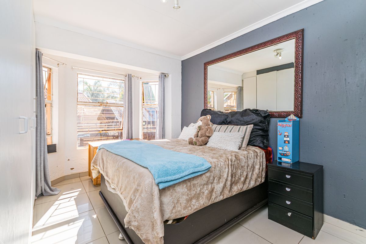 A bedroom featuring tiled flooring, built-in cupboards, and a bay window with security bars, offering a functional space with potential for cosmetic updates.