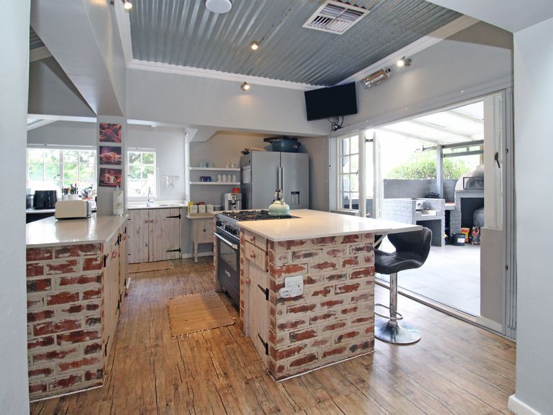 Open-plan kitchen featuring a rustic brick island, stone countertops, gas hob, and exposed corrugated metal ceiling, flowing seamlessly to an outdoor patio area.