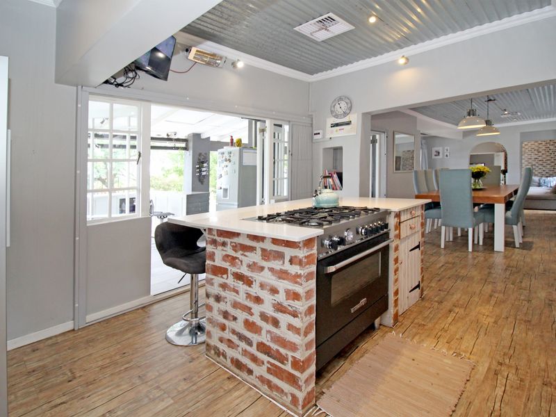 Open-plan kitchen and dining area featuring a brick-base island with gas stove, exposed corrugated metal ceilings, and timber flooring leading to an outdoor patio.