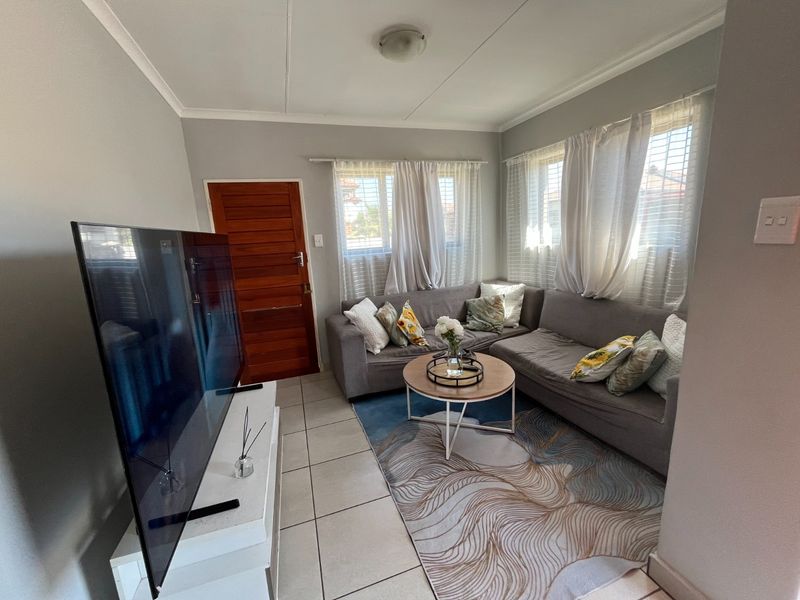 A compact living room featuring tiled flooring, a timber entrance door, and natural light through multiple windows with venetian blinds.