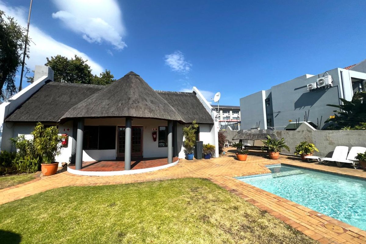 Rear exterior view featuring a thatched roof dwelling, covered patio, swimming pool, and paved garden area with boundary wall and electric fencing.
