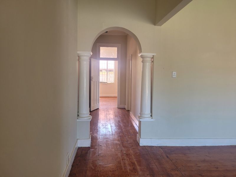 A bright, empty hallway featuring classic architectural archways with decorative columns and original hardwood flooring, leading towards a sunlit room.