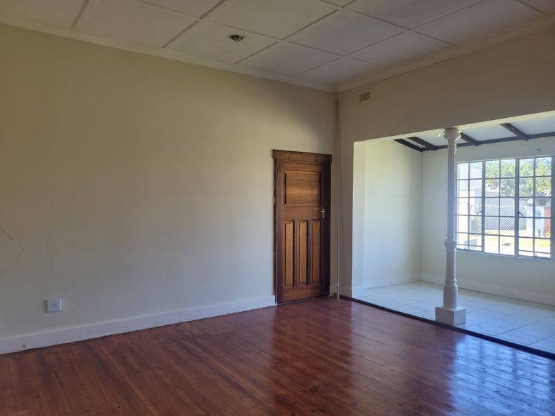 An empty living room featuring polished timber flooring, a classic timber door, and an adjoining sunroom area with tiled flooring and a support column.