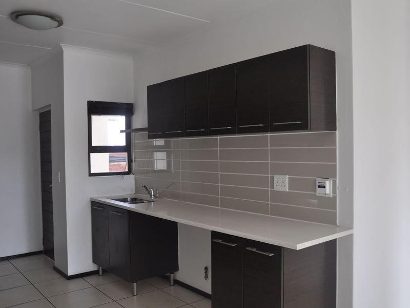A modern kitchen featuring dark wood cabinetry, light stone countertops, a stainless steel sink, and neutral tiled backsplash in an open-plan layout.