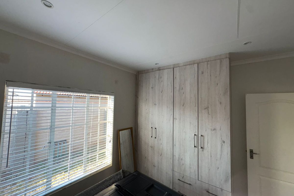 A bedroom featuring light wood-grain built-in cupboards, recessed ceiling lighting, and venetian blinds on the window.