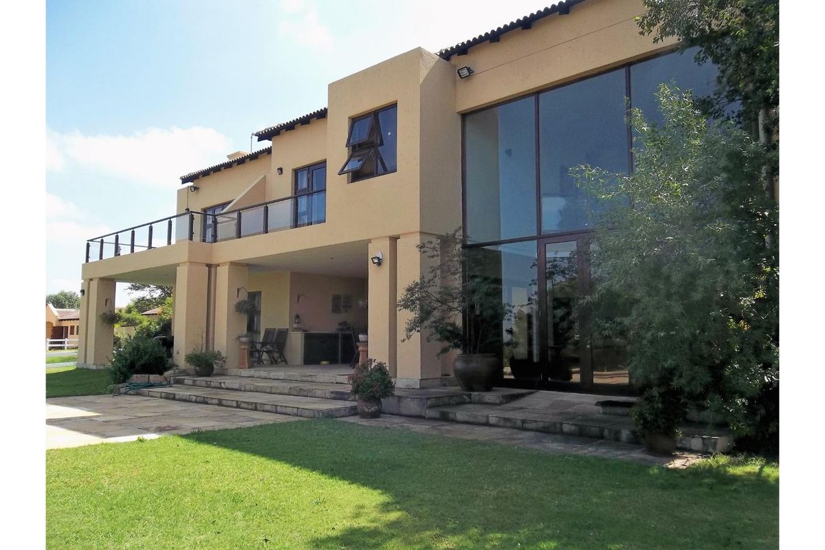 Rear view of a multi-story residence featuring a covered patio, expansive glass windows, and a balcony with glass balustrades overlooking the garden.