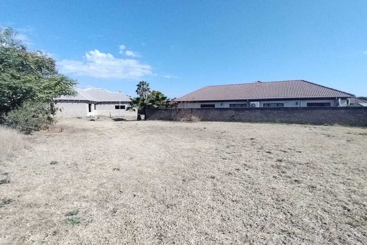 A vacant residential plot featuring dry grass, enclosed by a boundary wall with neighbouring properties visible in the background.