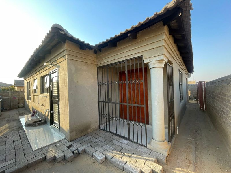 Exterior view of the house entrance featuring a covered porch with a decorative column and security gate, with unfinished paving in the foreground.