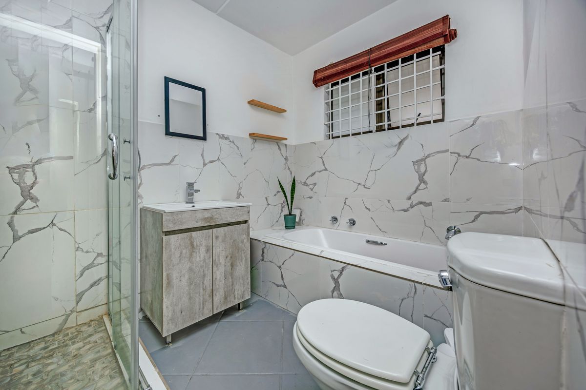 Modernized bathroom featuring a glass shower enclosure, built-in bathtub, vanity cabinet, and marble-look wall tiling with security bars on the window.
