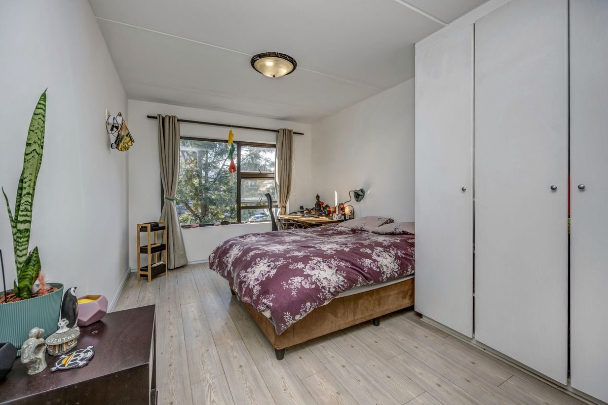A functional bedroom featuring light-toned laminate flooring, a large built-in wardrobe, and a window providing natural light.