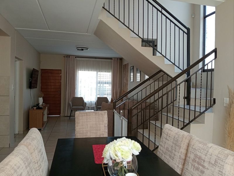 An open-plan living and dining area featuring a prominent staircase with a metal balustrade, tiled flooring, and natural light from large windows.