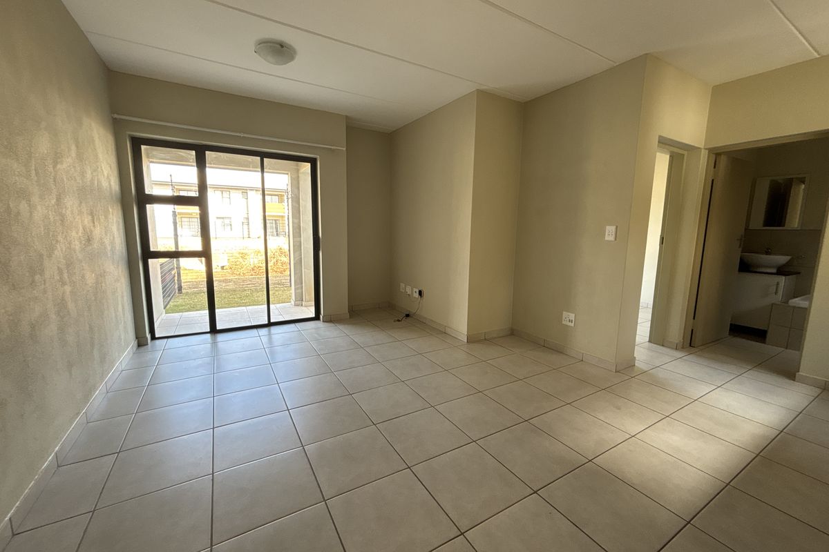 An empty, neutral-toned living room featuring ceramic floor tiles and a sliding glass door providing access to an outdoor patio area.