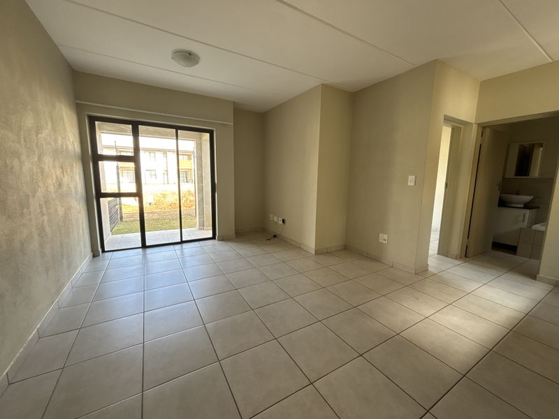An empty, neutral-toned living room featuring ceramic floor tiles and a sliding glass door providing access to an outdoor patio area.