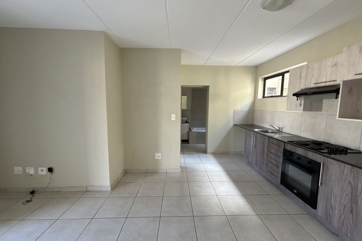 A clean, functional kitchen featuring modern wood-look cabinetry, built-in oven and hob, tiled flooring, and a neutral backsplash, leading into an adjacent bathroom area.