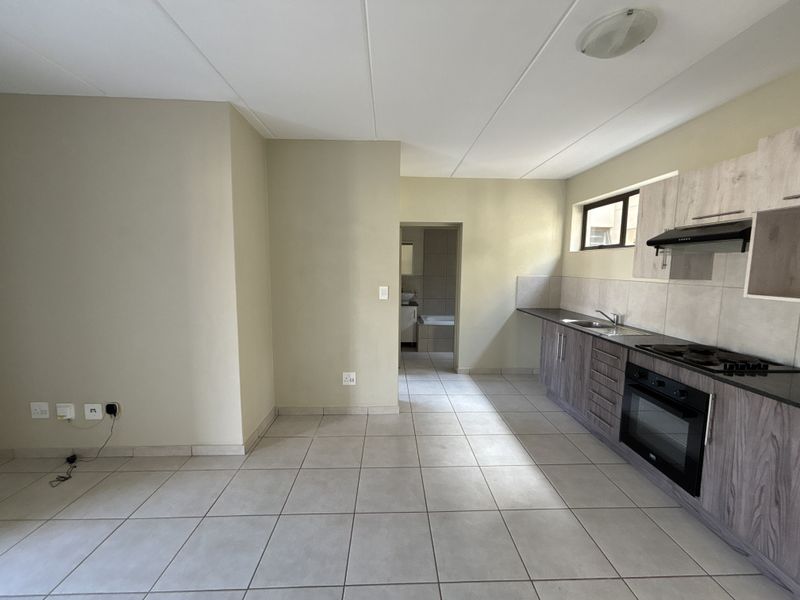 A clean, functional kitchen featuring modern wood-look cabinetry, built-in oven and hob, tiled flooring, and a neutral backsplash, leading into an adjacent bathroom area.