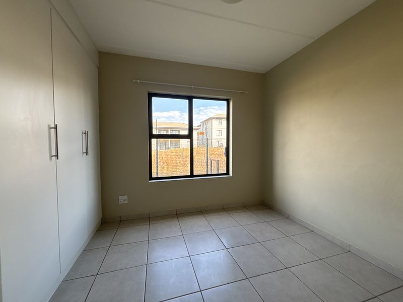 A clean, empty bedroom featuring neutral tiled flooring, built-in cupboards, and an aluminium-framed window providing natural light.