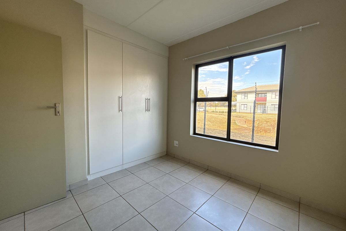 A bright, empty bedroom featuring neutral tiled flooring, built-in cupboards, and a large window providing natural light.