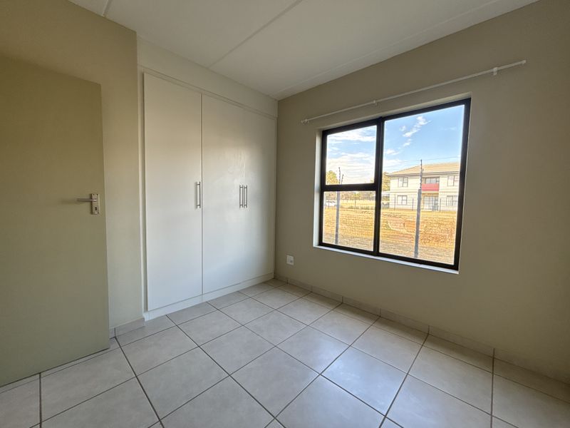 A bright, empty bedroom featuring neutral tiled flooring, built-in cupboards, and a large window providing natural light.