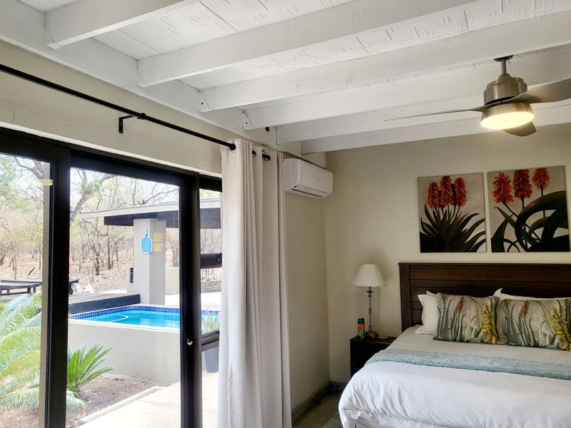 A well-maintained bedroom featuring exposed timber ceiling beams, a ceiling fan, air conditioning, and direct access to a private patio and pool area.