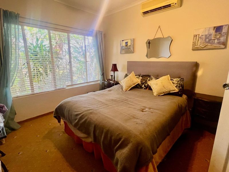 A bedroom featuring a large window with security bars, carpet flooring, and a wall-mounted split-system air conditioner.