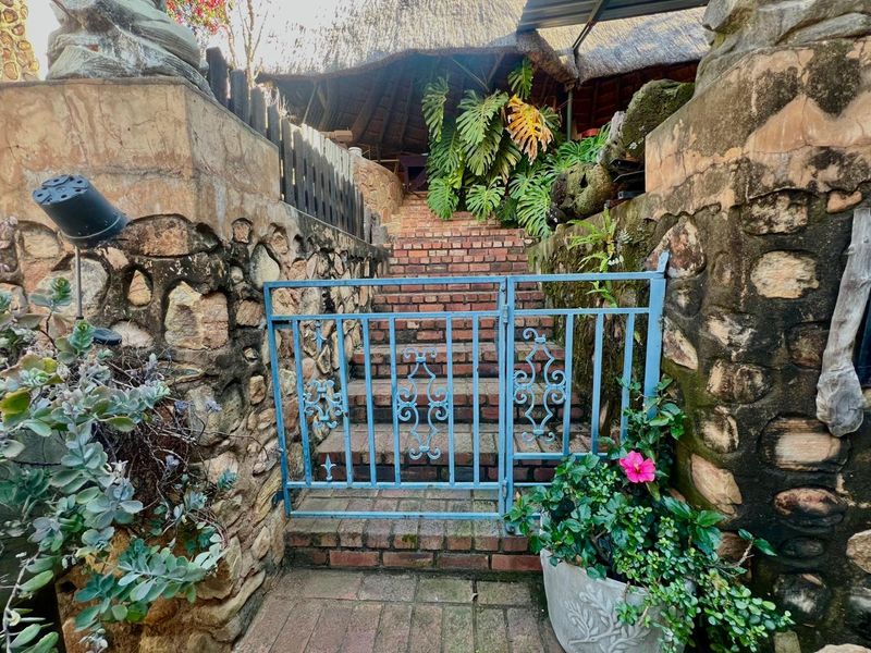 A rustic entrance featuring a brick staircase, stone retaining walls, and a wrought iron gate leading to a thatched roof structure.