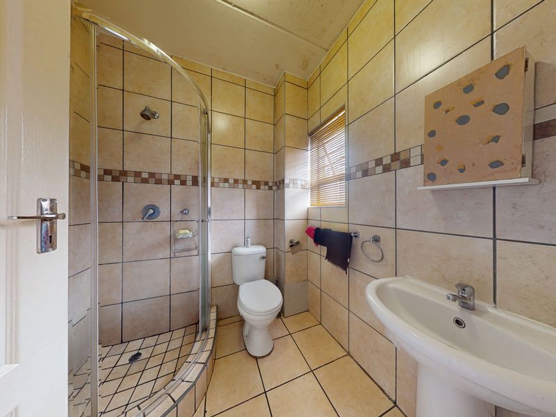 A compact bathroom featuring a curved glass shower enclosure, pedestal washbasin, and toilet, finished with neutral ceramic wall and floor tiling.