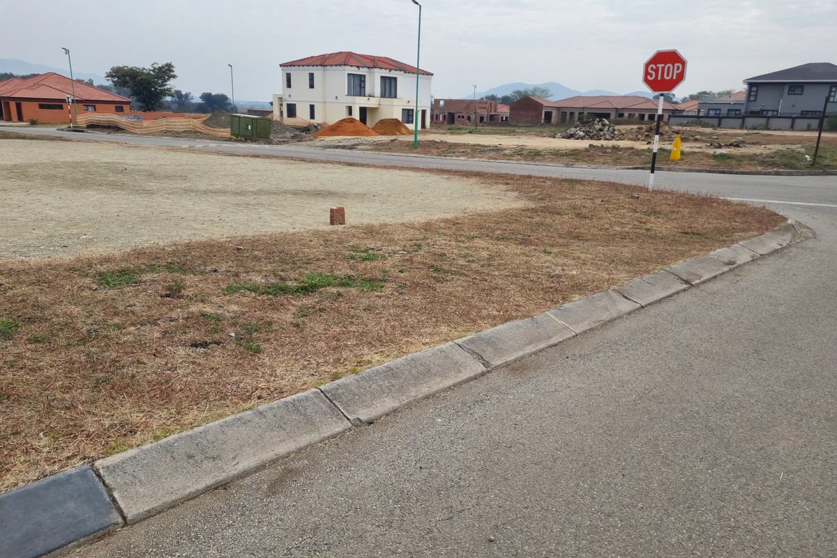 A street-level view of a vacant residential plot featuring paved road access, concrete curbing, and surrounding neighborhood development.