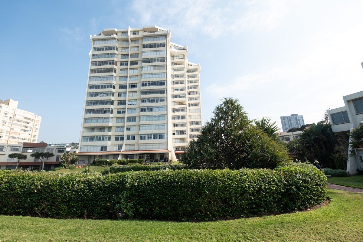 Exterior view of a multi-story apartment building featuring landscaped gardens and manicured hedges in the foreground.