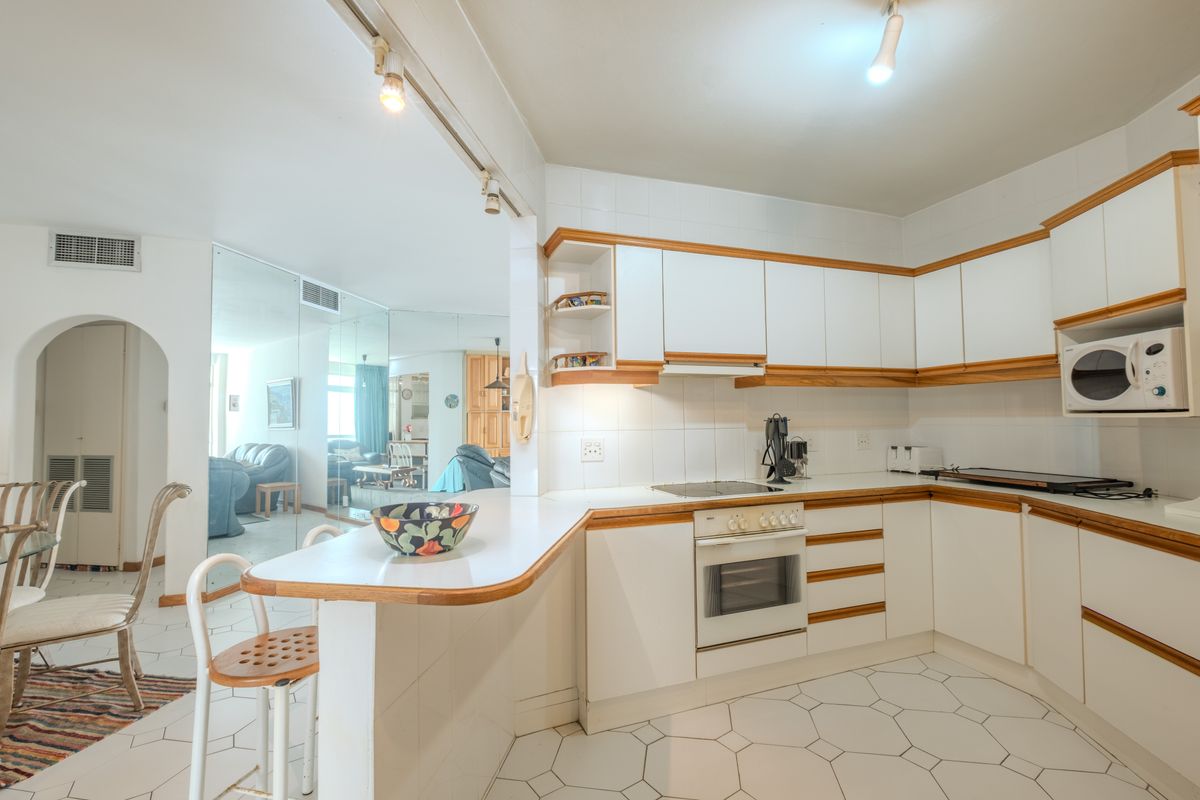 A functional kitchen featuring white cabinetry with timber trim, built-in oven, hob, and a breakfast bar, opening into the adjacent living area.
