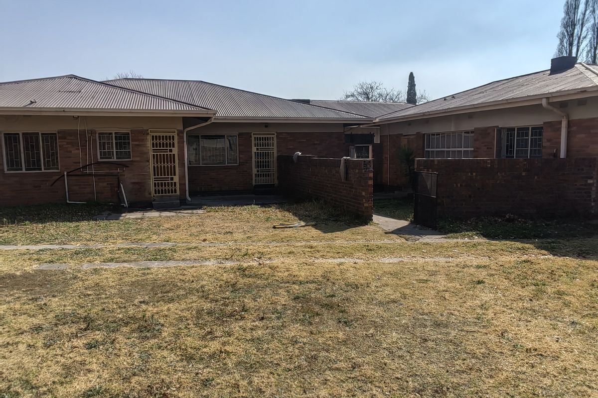 Rear view of a single-story brick house featuring a corrugated metal roof, security gates, and a large backyard area with a paved walkway.