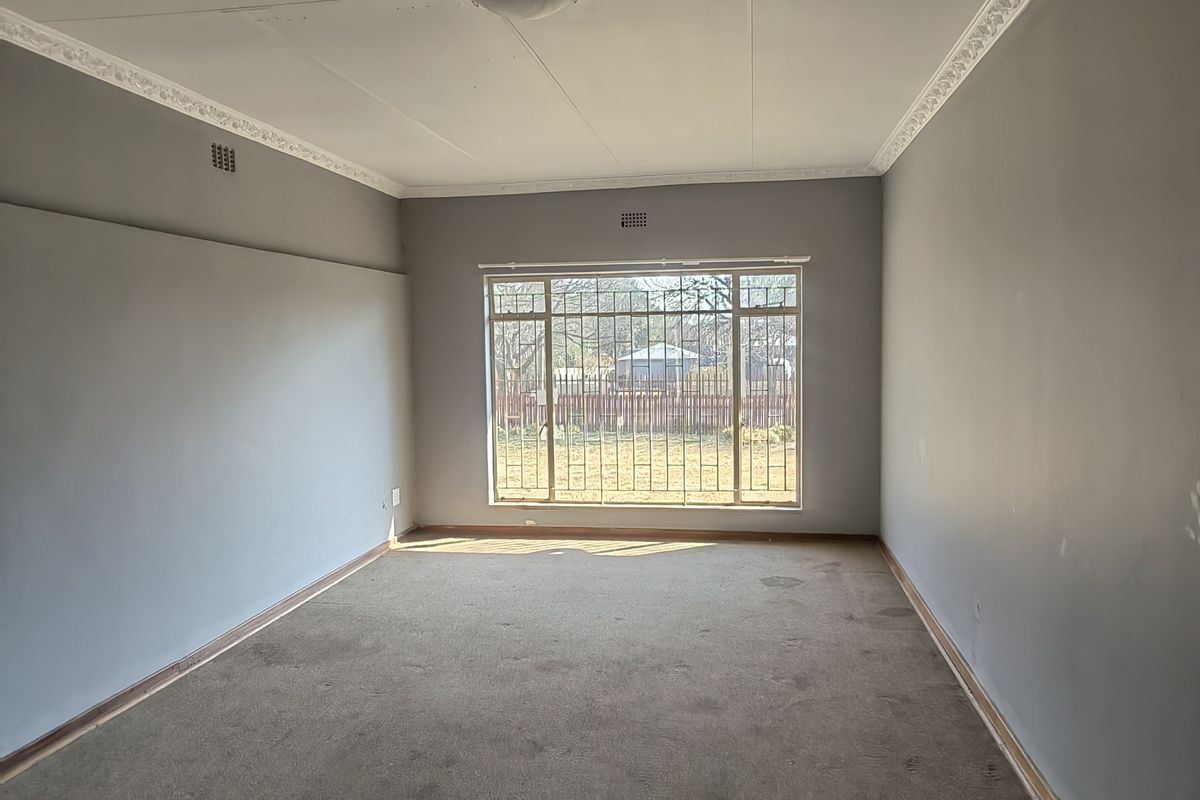 An empty living room featuring grey walls, decorative ceiling cornices, and a large window with security bars, offering a blank canvas for renovation.