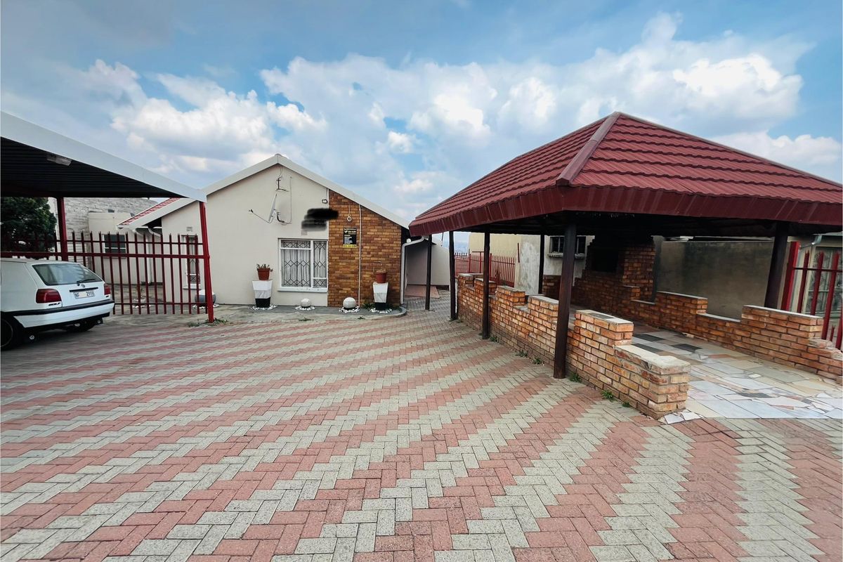 Exterior view of the property featuring a paved driveway, a covered carport, and a brick-built outdoor entertainment area with a tiled roof.