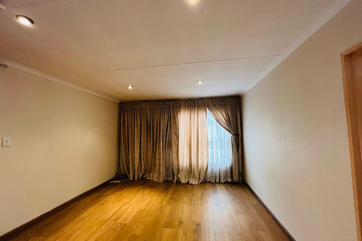 A bright, empty living room featuring warm-toned laminate wood flooring, recessed ceiling lighting, and full-length window curtains.
